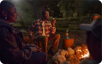 Friends are camping near a fire in the woods at night while a man is placing a marshmallow on a stick.
