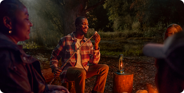 Friends are camping near a fire in the woods at night while a man is placing a marshmallow on a stick.